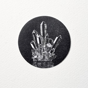 May include: A black and white illustration of a cluster of crystals. The crystals are depicted with sharp, angular facets and are arranged in a dense, upright formation within a black circular background. The image is set against a white backdrop.
