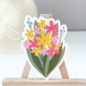 May include: A sticker featuring a bouquet of pink, yellow, and blue flowers with green leaves. The sticker is on a small wooden easel.