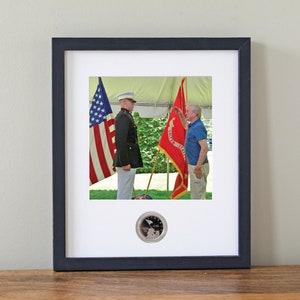 May include: A framed photograph of a man in a military uniform saluting another man in a casual outfit. The photo is taken outdoors with an American flag and a red flag with the words "United States" visible in the background. A silver coin is displayed in the lower center of the frame.