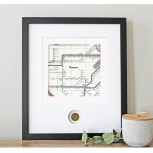 May include: A framed print of a vintage New York City subway map with the word "Queens" highlighted. The map is in black, white, and shades of blue, red, and brown. A small, round, gold-colored token is visible in the lower right corner of the frame.