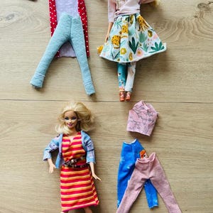 Könnte beinhalten: Eine Sammlung von Puppenkleidung, darunter eine rot-lila gemusterte Jacke, hellblaue Hosen, ein rosa Oberteil, ein Blumenrock und ein gestreiftes Kleid. Zwei Puppen tragen einige der Kleidungsstücke. Die Kleidung ist handgefertigt.