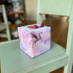 Könnte beinhalten: Ein rosa-lilafarbener Stoffwürfel mit einem Einhorn- und Flügelmotiv. Der Würfel hat zwei lilafarbene Stoffohren oben drauf.