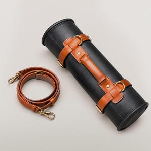 Könnte beinhalten: Ein zylindrisches schwarzes Etui mit hellbraunen Lederriemen, einem Tragegriff und goldfarbenen Metallbeschlägen. Das Etui verfügt über zwei umlaufende hellbraune Lederbänder mit D-Ringen und einen abnehmbaren, verstellbaren Schulterriemen.