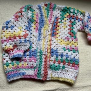 May include: A colourful crocheted cardigan with a button-up front. The cardigan is made with a variety of pastel colours, including pink, blue, yellow, and green.