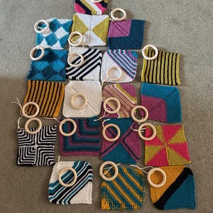 May include: A collection of square knitted fabric swatches in various colours and patterns, including stripes, diamonds, and geometric blocks. Each individual square features a small, light-coloured wooden ring attached by a thin cord.