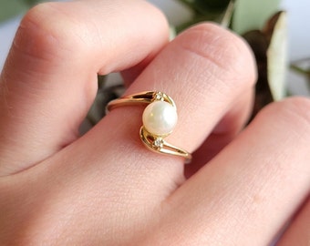 May include: A gold ring with a single pearl and two small diamonds. The ring is on a finger.