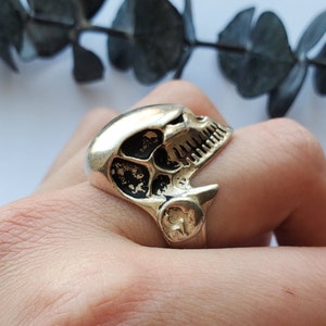 May include: A silver skull ring with a detailed design. The ring is worn on a person's finger.
