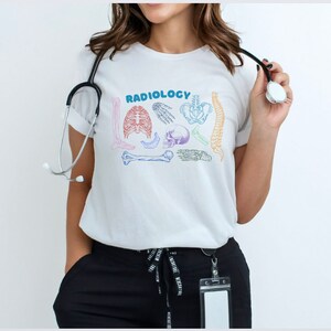 Op de afbeelding: Wit T-shirt met het woord "RADIOLOGY" in regenboogkleuren en een afbeelding van verschillende botten en een schedel.