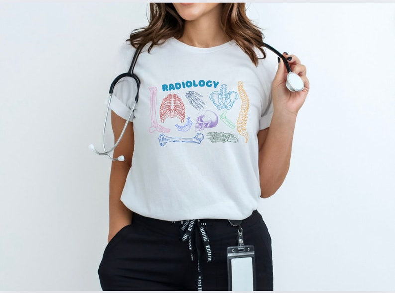 Op de afbeelding: Wit T-shirt met het woord "RADIOLOGY" in regenboogkleuren en een afbeelding van verschillende botten en een schedel.