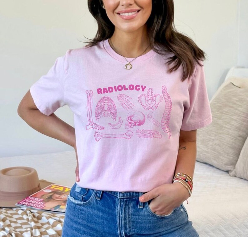 Op de afbeelding: Een roze T-shirt met het woord "RADIOLOGY" in roze op de voorkant. Het shirt heeft een afbeelding van verschillende botten en een schedel.