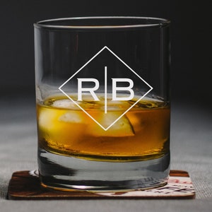 May include: A clear glass with a diamond-shaped design etched with the initials "R B" and filled with amber liquid and ice cubes. The glass is sitting on a brown coaster.