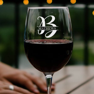 May include: A clear wine glass with a personalized design. The glass has the letter "B" and the name "Mathews" etched on it. The glass is filled with red wine.