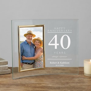 May include: A clear glass photo frame with a gold border, featuring a photo of a couple and the text "Happy Anniversary 40 Years". The frame is displayed on a wooden surface.