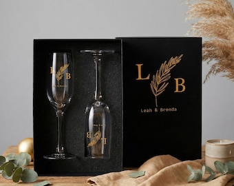 May include: A gift set featuring two champagne flutes and a black box. The flutes have gold lettering and a wheat design. The box has gold lettering that reads "L B" with a wheat design and the names "Leah & Brenda".