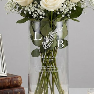 May include: A bouquet of white roses and baby's breath in a clear glass vase. The vase has the initials "L B" and a decorative leaf design. A silver picture frame, books, and a candle are also present.