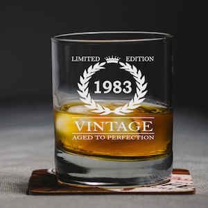 May include: A clear glass tumbler with the text "Limited Edition 1983 Vintage Aged to Perfection" etched on the side. The glass is filled with amber liquid and ice cubes.