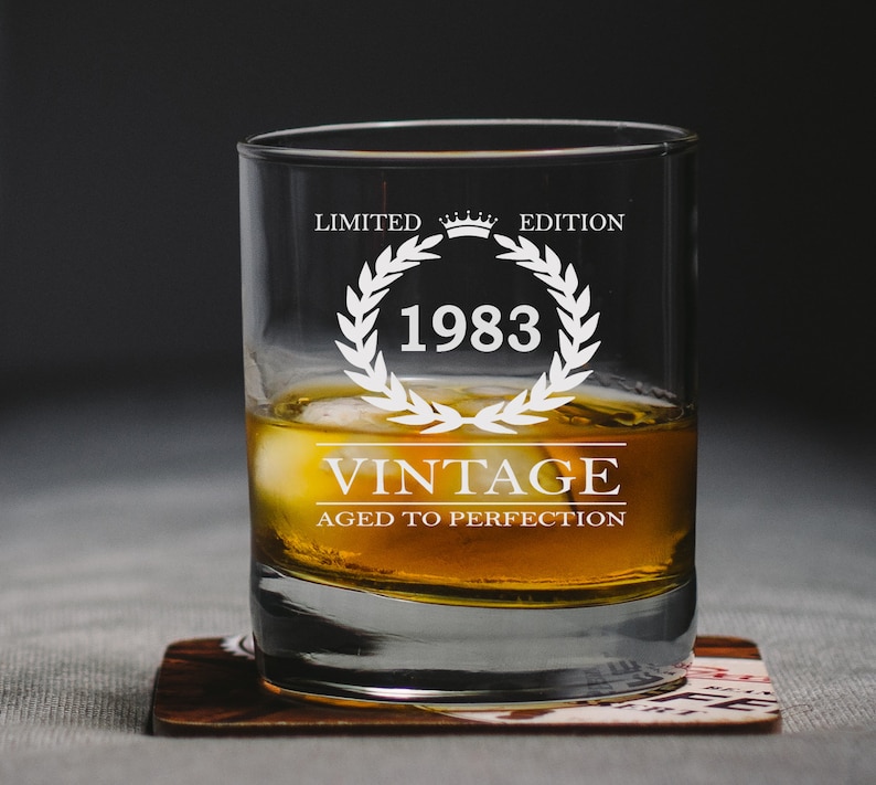 May include: A clear glass tumbler with the text "Limited Edition 1983 Vintage Aged to Perfection" etched on the side. The glass is filled with amber liquid and ice cubes.