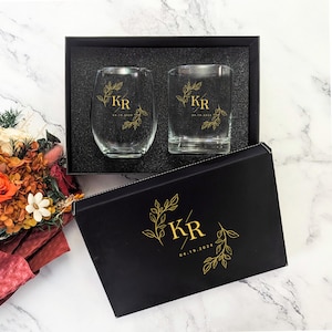 May include: A gift set featuring two clear drinking glasses, one stemless and one with a flat base, inside a black box. Each glass is adorned with gold-colored initials and a date. The box also has matching gold leaf designs.