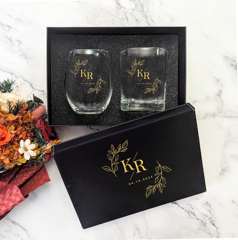 May include: A gift set featuring two clear drinking glasses, one stemless and one with a flat base, inside a black box. Each glass is adorned with gold-colored initials and a date. The box also has matching gold leaf designs.