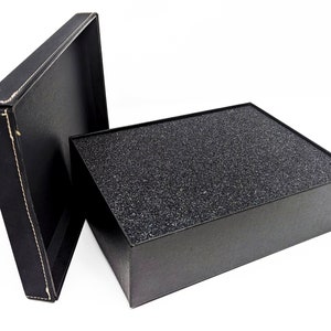 May include: An open black gift box with a textured, dark gray interior and a separate lid. The lid is black with white stitching around the edges. The box is on a white background.