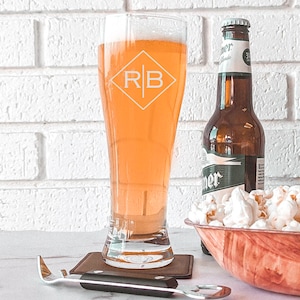 Pode incluir: Um copo de cerveja personalizado com as iniciais "RB" gravadas em forma de diamante. O copo est&aacute; cheio de cerveja de cor clara e est&aacute; sobre um descanso de copo castanho. No fundo, h&aacute; uma garrafa de cerveja e uma tigela de pipocas.