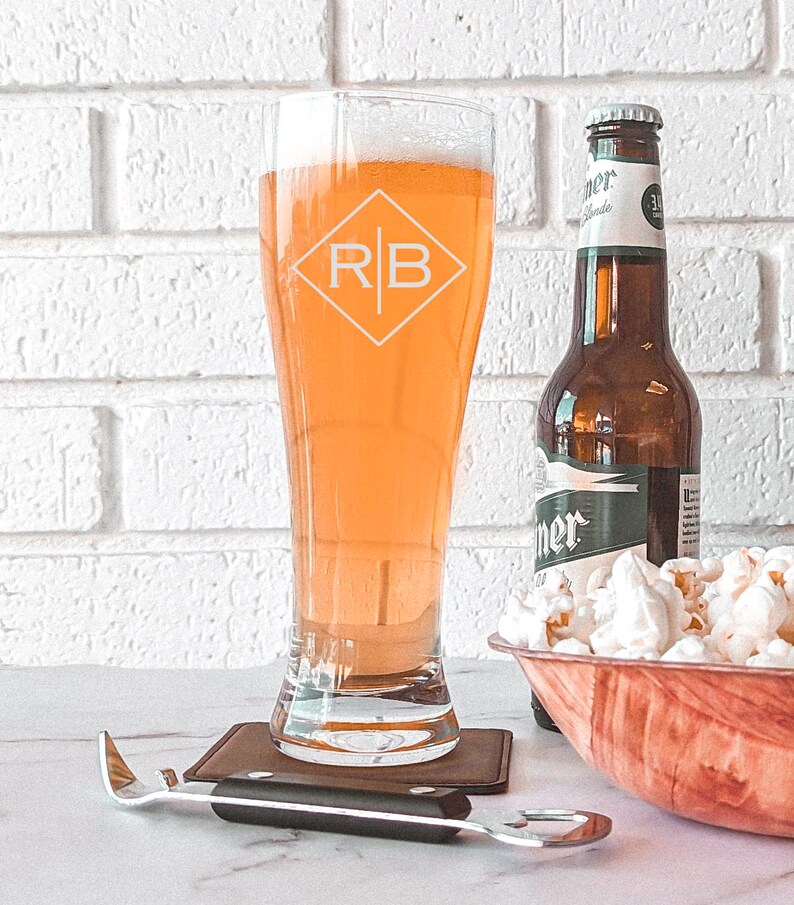 Pode incluir: Um copo de cerveja personalizado com as iniciais "RB" gravadas em forma de diamante. O copo est&aacute; cheio de cerveja de cor clara e est&aacute; sobre um descanso de copo castanho. No fundo, h&aacute; uma garrafa de cerveja e uma tigela de pipocas.