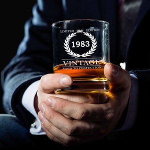 May include: A clear glass whiskey tumbler with the text "Limited Edition 1983 Vintage Aged to Perfection" etched on the front. The glass is filled with amber liquid and is being held by a person's hand.