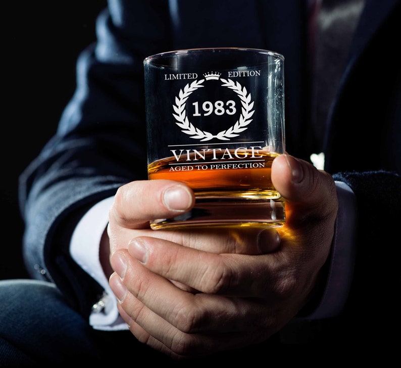 May include: A clear glass whiskey tumbler with the text "Limited Edition 1983 Vintage Aged to Perfection" etched on the front. The glass is filled with amber liquid and is being held by a person's hand.