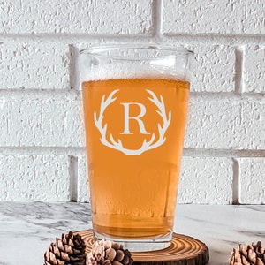 Pode incluir: Uma caneca de cerveja de vidro transparente com um design de chifre de veado branco e a letra "R" no centro. A caneca est&aacute; cheia de uma cerveja &acirc;mbar clara e est&aacute; sobre um descanso de copo de madeira.