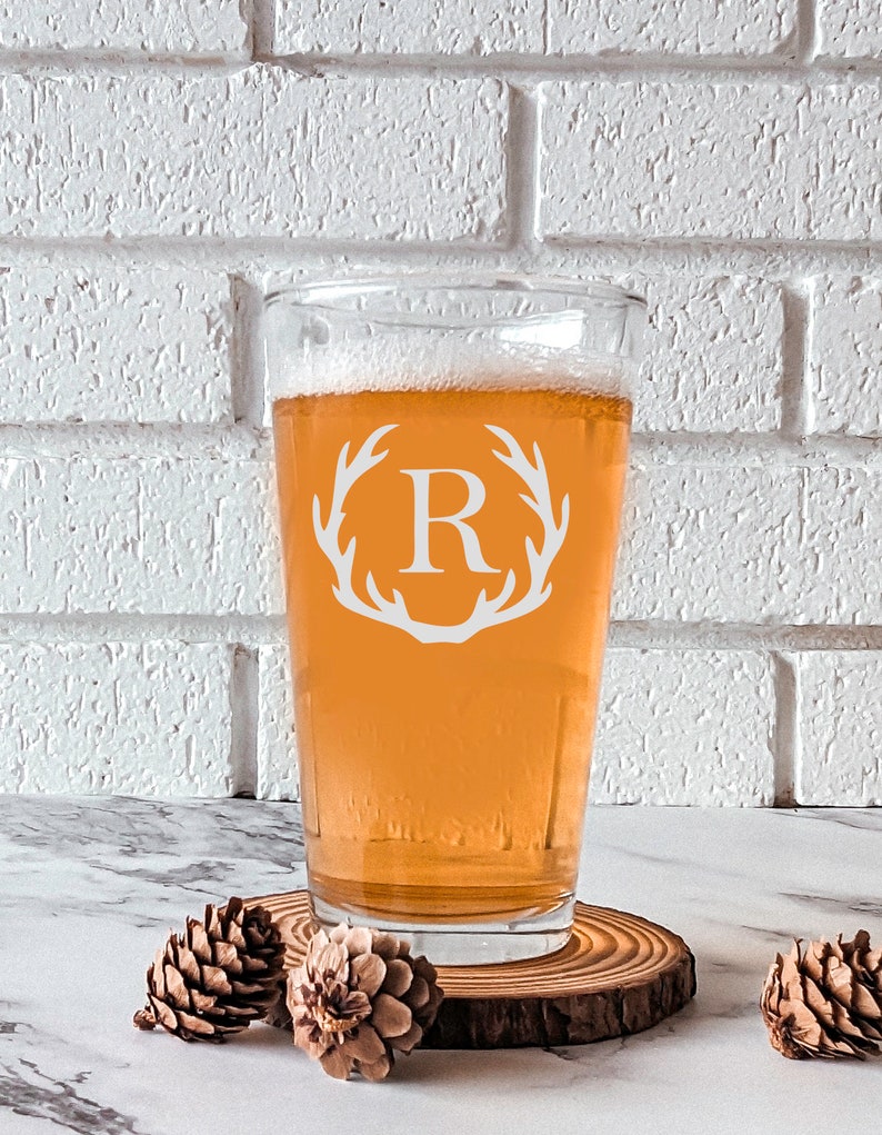 Pode incluir: Uma caneca de cerveja de vidro transparente com um design de chifre de veado branco e a letra "R" no centro. A caneca est&aacute; cheia de uma cerveja &acirc;mbar clara e est&aacute; sobre um descanso de copo de madeira.
