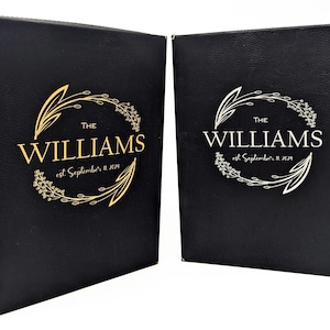 May include: Two black leather-bound books with gold and silver foil lettering. The text reads "THE WILLIAMS" with "est. September 11, 2019" below. A floral wreath design surrounds the text. The books are likely for a wedding or special event.
