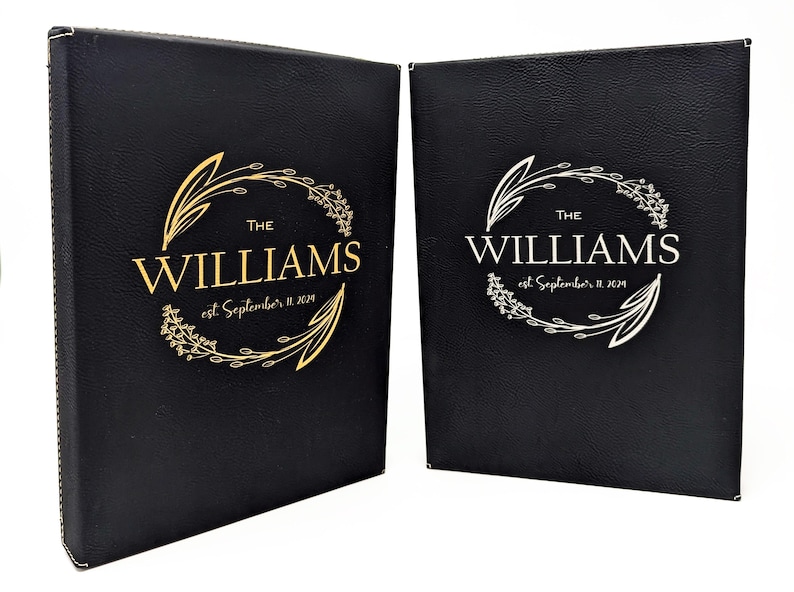 May include: Two black leather-bound books with gold and silver foil lettering. The text reads "THE WILLIAMS" with "est. September 11, 2019" below. A floral wreath design surrounds the text. The books are likely for a wedding or special event.