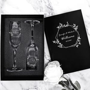 May include: Two engraved champagne flutes in a black box with a white rose. The flutes are engraved with a geometric design and the text "Jennifer & Michael Williams 08.15.2025".