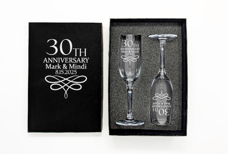 May include: A black gift box with the text "30th Anniversary Mark & Mindi 8.15.2025" next to a box containing two champagne flutes. The flutes are engraved with the same text and decorative flourishes.