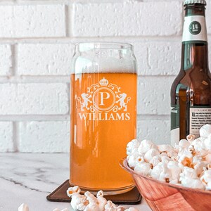 Pode incluir: Um copo de cerveja de vidro personalizado com um bras&atilde;o que apresenta a letra "P" e o nome "WILLIAMS" gravado no lado. O copo est&aacute; cheio de uma cerveja de cor clara e est&aacute; sobre um descanso de copo castanho. H&aacute; uma tigela de pipocas no fundo.