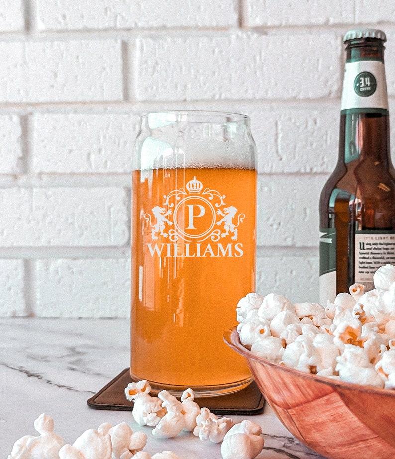 Pode incluir: Um copo de cerveja de vidro personalizado com um bras&atilde;o que apresenta a letra "P" e o nome "WILLIAMS" gravado no lado. O copo est&aacute; cheio de uma cerveja de cor clara e est&aacute; sobre um descanso de copo castanho. H&aacute; uma tigela de pipocas no fundo.