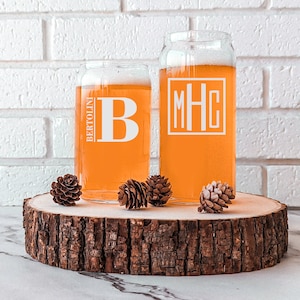 Pode incluir: Duas canecas de cerveja de vidro transparente com monogramas personalizados. A caneca &agrave; esquerda tem a letra "B" e o texto "BERTOLINI". A caneca &agrave; direita tem o monograma "MHC". As canecas est&atilde;o sobre uma fatia de madeira.