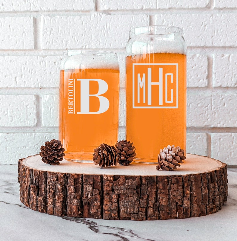 Pode incluir: Duas canecas de cerveja de vidro transparente com monogramas personalizados. A caneca &agrave; esquerda tem a letra "B" e o texto "BERTOLINI". A caneca &agrave; direita tem o monograma "MHC". As canecas est&atilde;o sobre uma fatia de madeira.