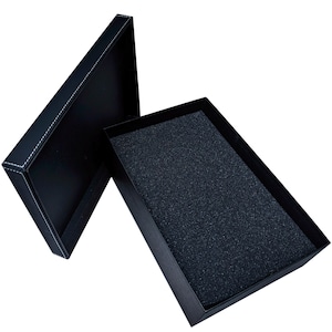 May include: Black rectangular gift box with a lid, open to reveal a dark interior. The lid is slightly ajar, showcasing white stitching along the edges. The inside of the box has a textured, dark gray foam lining.
