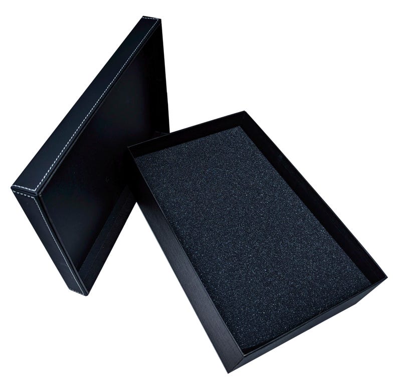 May include: Black rectangular gift box with a lid, open to reveal a dark interior. The lid is slightly ajar, showcasing white stitching along the edges. The inside of the box has a textured, dark gray foam lining.
