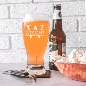 Pode incluir: Um copo de cerveja personalizado com o nome "WRIGHT" gravado nele, ao lado de uma garrafa de cerveja e uma tigela de pipoca. O copo est&aacute; sobre um descanso de copo castanho.