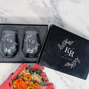 May include: Two stemless wine glasses with the initials "KR" and a floral design, presented in a black gift box. The box also features the initials and date "06.15.2026". A bouquet of flowers is in the foreground.