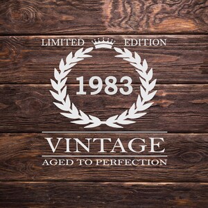 May include: White text on a brown wood background reads "Limited Edition" with a crown symbol above the text. A white laurel wreath surrounds the year "1983". Below the wreath, the text reads "Vintage Aged to Perfection".