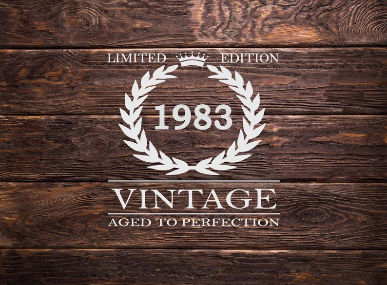May include: White text on a brown wood background reads "Limited Edition" with a crown symbol above the text. A white laurel wreath surrounds the year "1983". Below the wreath, the text reads "Vintage Aged to Perfection".