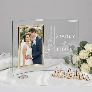 May include: A clear glass photo frame with a gold-colored border, displaying a wedding photo. The frame is engraved with "Happy Ever After" and the names "Brandy & Loren" with a date. A wooden "Mr & Mrs" sign is at the base.