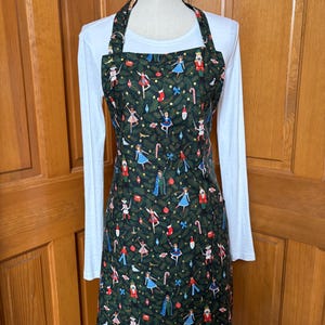 May include: A dark green apron featuring a repeating pattern of nutcrackers, ballerinas, candy canes, and Christmas ornaments. The apron has a square neckline, adjustable neck straps with metal hardware, and a full-length design covering the torso.