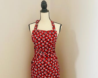 May include: A red apron with a black and white snowflake pattern. The apron is displayed on a mannequin and has a halter neck and a pocket on the right side. The apron is made of fabric and is designed for kitchen use.