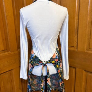 Peut inclure: Tablier &agrave; imprim&eacute; floral sur fond sombre, avec des fleurs roses, bleues et jaunes. Il est nou&eacute; &agrave; la taille et poss&egrave;de un col halter. Le tablier est port&eacute; sur un haut blanc &agrave; manches longues.