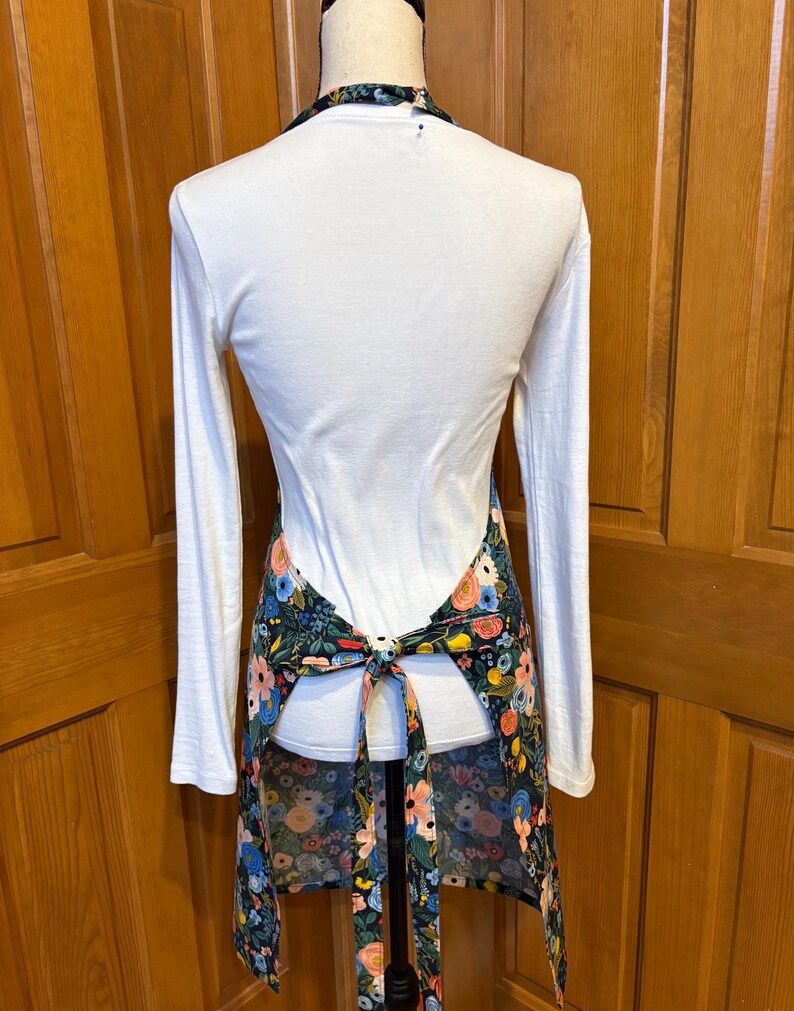 Peut inclure: Tablier &agrave; imprim&eacute; floral sur fond sombre, avec des fleurs roses, bleues et jaunes. Il est nou&eacute; &agrave; la taille et poss&egrave;de un col halter. Le tablier est port&eacute; sur un haut blanc &agrave; manches longues.