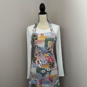 May include: A patchwork apron with a variety of floral and geometric patterns in shades of red, blue, yellow, and green. The apron is displayed on a mannequin, showcasing its halter neck and waist tie design. The apron is a kitchen or craft accessory.